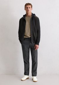 Junger Mann steht vor einer weißen Wand und trägt ein beiges Hemd, einen dunklen Reißverschluss-Hoodie, graue Hose und weiße Sneakers.