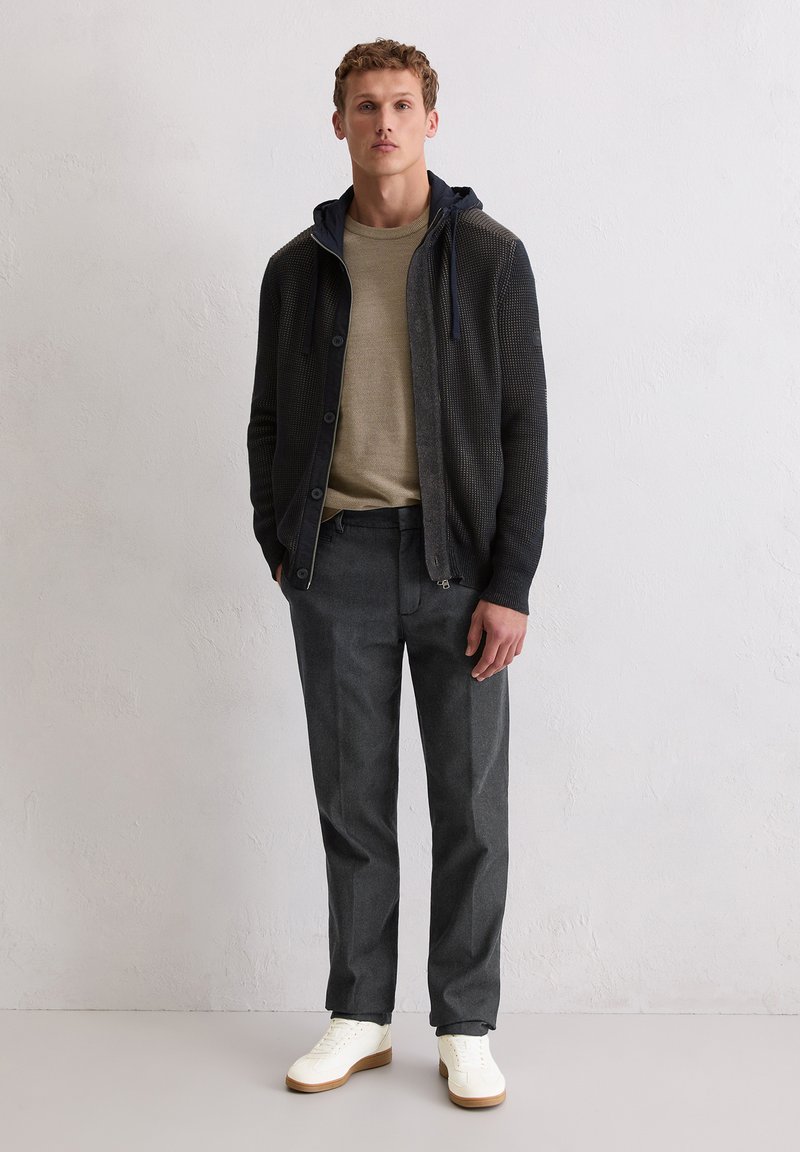 Junger Mann steht vor einer weißen Wand und trägt ein beiges Hemd, einen dunklen Reißverschluss-Hoodie, graue Hose und weiße Sneakers.