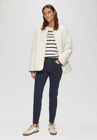 Veste crème matelassée, pull rayé noir et blanc, pantalon ajusté bleu marine, baskets beige et gris, debout sur un fond neutre.