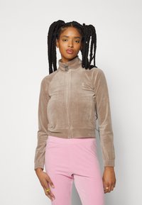 Braune Velourjacke mit Reißverschluss, hohem Kragen und seitlichen Taschen, kombiniert mit hellrosa, eng anliegenden Hosen. Sichtbare Details aus silbernem Metall.