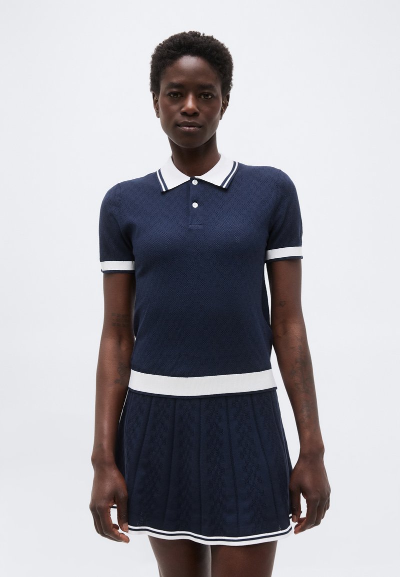 Γυναίκα με κοντά σγουρά μαλλιά που φοράει μπλε πλεκτό πουλόβερ πόλο και ταιριαστή πλισέ φούστα με λευκή διακόσμηση, στέκεται μπροστά σε απλό φόντο.