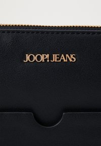 Čierna kožená peňaženka s textúrovaným povrchom, s zlatým logom "JOOP! JEANS" a zipsom na vrchu.