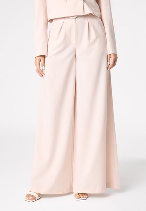 Pantalons larges roses avec des plis, fabriqués à partir d'un tissu lisse. Présente une taille haute et une silhouette fluide, associés à des sandales blanches.