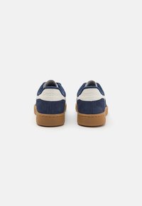 Kék velúr sportcipők fehér kiegészítőkkel, texturált gumi talppal és "Reebok" felirattal a sarokrészen. Lekerekített orrú kialakítás.