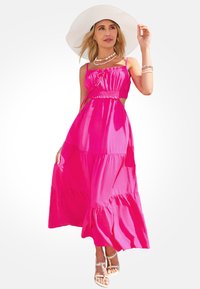 Robe maxi en rose vif, avec des bretelles spaghetti, un bustier ajusté, des volants pour du volume et un tissu léger. Complétée par un chapeau à large bord.