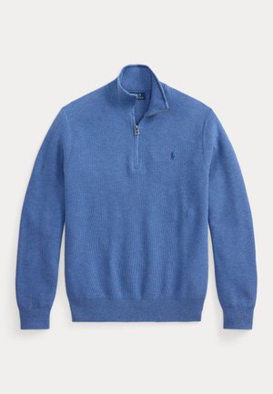 Homme portant un pull à zip court bleu par-dessus une chemise à col, pantalon noir et mocassins marron, debout les mains dans les poches regardant sur le côté.