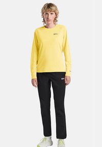 Langärmliges gelbes Hemd mit einem Logo auf der Brust, kombiniert mit schwarzen Hosen. Das Hemd hat ein schlichtes Design; die Hose ist gerade geschnitten.