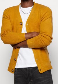 Hořčicově žlutý kardigan z texturované látky, s žebrovanými manžetami, výstřihem do V a pěti hnědými knoflíky vpředu.