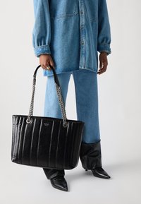 Zwarte leren tote bag met verticale geribbelde textuur, zilveren kettinghandvatten en een logo plaatje. gedragen door iemand in denimkleding.