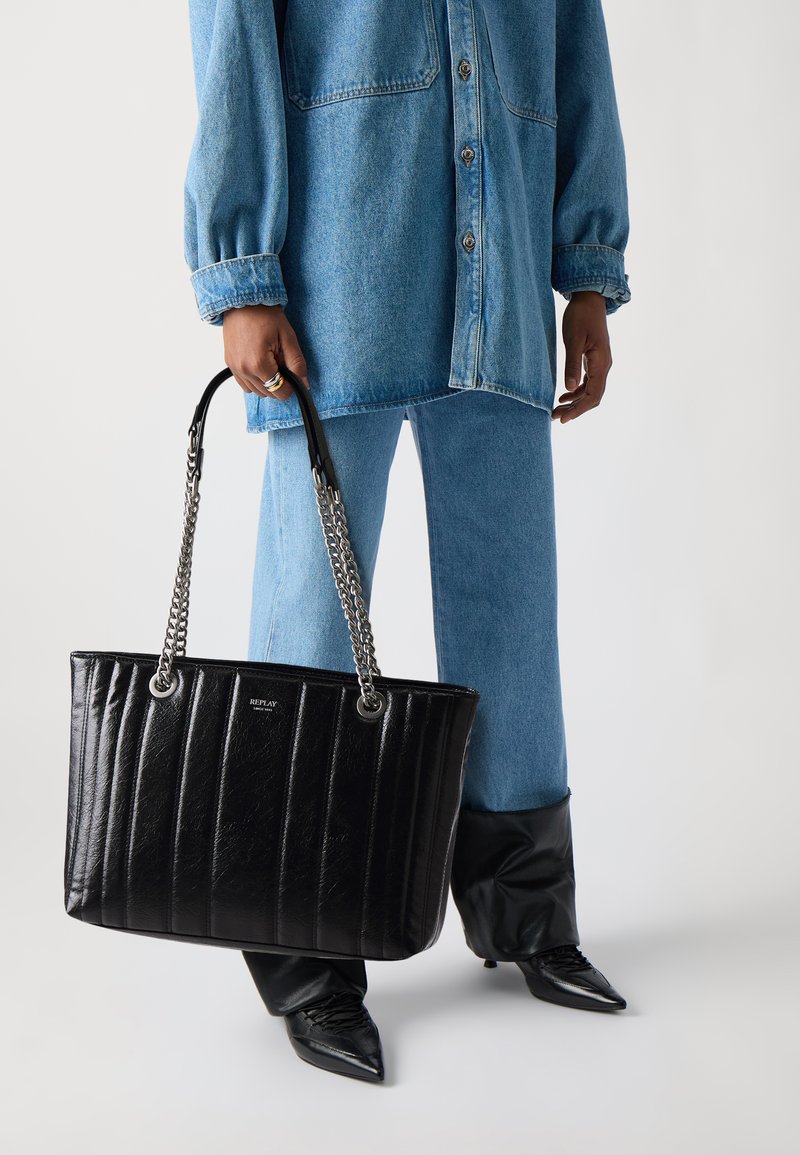 Zwarte leren tote bag met verticale geribbelde textuur, zilveren kettinghandvatten en een logo plaatje. gedragen door iemand in denimkleding.