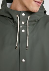 Zbliżenie na osobę w ciemnozielonym płaszczu przeciwdeszczowym z kapturem, białymi sznurkami ściągającymi i srebrnymi zatrzaskami.