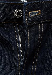 Tmavé džíny z denimu s kovovým knoflíkem a zipem. Obsahují kontrastní žluté prošívání a malou poutko na opasek blízko zapínání.