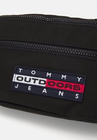 Bolsa de tela negra con cierres de doble cremallera. Logotipo bordado destacado con "TOMMY" y "OUTDOORS" en colores rojo, azul marino y blanco.