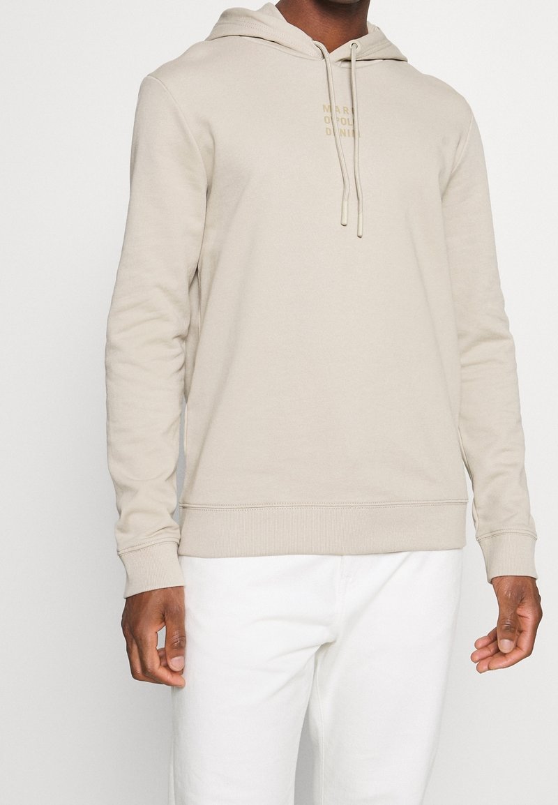 Homme portant un sweat à capuche beige clair avec des cordons et un texte discret sur la poitrine, accompagné d'un pantalon blanc, posant devant un fond uni.