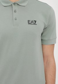 Svetlozelená polo košeľa vyrobená z jemného materiálu, s tri gombíkmi na zapínanie, krátkymi rukávmi a čiernym logom na ľavej hrudi.