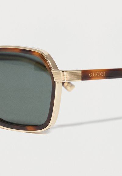 Κοντινή λήψη γυαλιών ηλίου με χρυσό πλαίσιο και σκουρόχρωμους φακούς, με μπράτσα χελώνας που φέρουν το λογότυπο της Gucci σε χρυσά γράμματα.