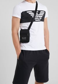 Borsa a tracolla nera in nylon con zip, caratterizzata da un emblema EA7. Indossata sopra una maglietta bianca con un design grafico nero e pantaloni corti scuri.