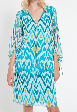 Vrouw draagt een knielange jurk met lange doorschijnende mouwen, met een blauw, wit en groen zigzag- en diamantpatroon.