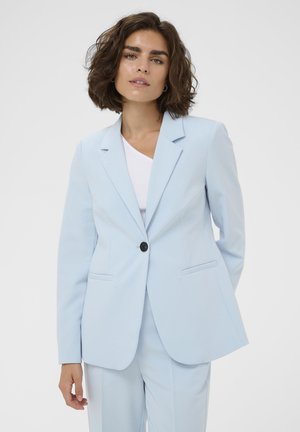 Kobieta z kręconymi brązowymi włosami w jasnoniebieskim żakiecie na jedno zapięcie i dopasowanych spodniach, stojąca na tle jednolitego tła, w białym topie.