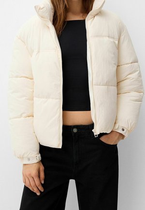 Giacca a vento in materiale bianco sporco, chiusura con zip, colletto alto, design trapuntato, con tasche laterali, indossata sopra un crop top nero e pantaloni neri.