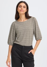Blouse marron à motifs avec des manches bouffantes et un col rond, assortie à un pantalon noir, présentée contre un fond clair.