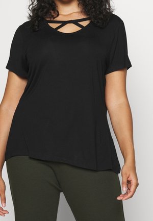 Femme portant un haut noir à manches courtes avec un détail de col croisé et un pantalon vert olive, vue des épaules aux hanches sur fond uni.