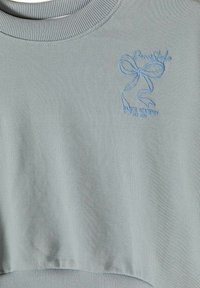 Sweat-shirt en maille bleu clair avec col et ourlet côtelés, arborant un design de nœud brodé et un texte portant "Dance Academy Est. 1978" sur la poitrine.