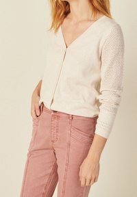 Ljust beige cardigan med V-halsdesign och stickade, mönstrade ärmar. Matchad med rosa denimbyxor med synlig sömnad och fickor.