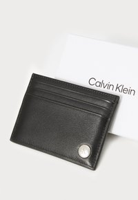 Crni kožni novčanik za kartice s više utora i srebrnim kružnim logotipom. Postavljen pored bijele kartice s natpisom "Calvin Klein".