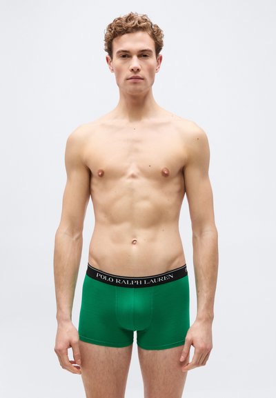 Νέος άνδρας μοντέλο στέκεται, φορώντας πράσινα εσώρουχα boxer Polo Ralph Lauren με μαύρη ζώνη, ουδέτερη έκφραση, απλό φωτεινό φόντο.