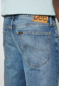 Jeans in denim blu con un lavaggio sbiadito, dotati di tasche posteriori curve. È visibile una patch in pelle color cuoio con il marchio "Lee" in vita.