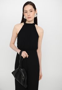 Svart halterneck topp med samlet midje, kombinert med en svart knyttet håndveske. Modellen har på seg sølvfargede statement øreringer og rett hår.