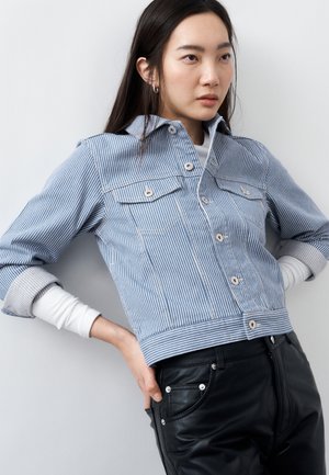 Νεαρή γυναίκα με ριγέ μπλε σακάκι και μαύρο δερμάτινο παντελόνι, ποζάροντας με τα χέρια στη μέση μπροστά σε ένα απλό φόντο.