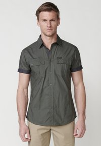 Camisa de manga corta verde oscuro con diseño de botones, que presenta dos bolsillos frontales, un cuello en contraste y una tela suave y texturizada.