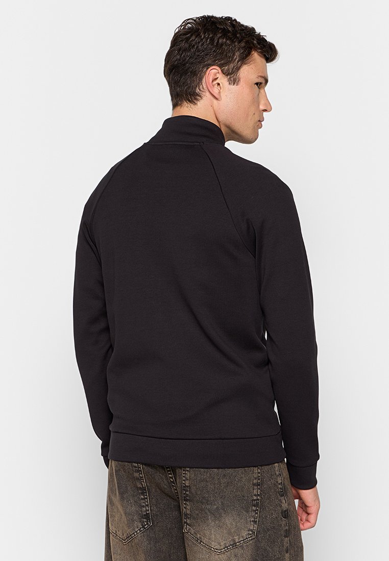 Uomo che indossa un maglione nero a maniche lunghe e jeans grigi scuri sbiaditi, mostrato di spalle contro uno sfondo chiaro e semplice.