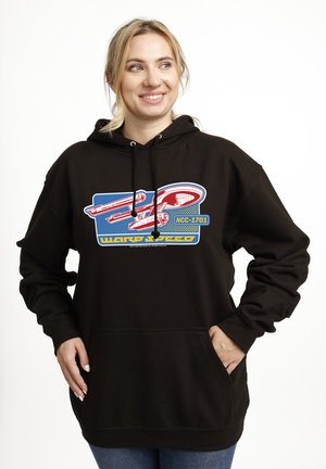 Kvinde smilende, iført sort hættetrøje med rød og blå stjerneskibsgrafik mærket "NCC-1701 WARP SPEED", hænder i forlommer.