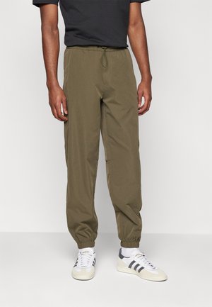Olivgrüne Jogginghosen mit einem elastischen Bund und Kordelzug. Mit Bündchen an den Knöcheln und einer glatten Textur. Getragen mit weißen Sneakers.