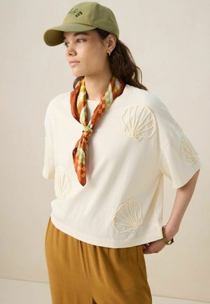 White cotton t-shirt with shell embroidery, paired with an orange patterned scarf and olive cap; brown trousers. Plain background.