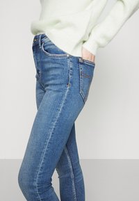 Jeans de denim azul de corte Skinny con cintura alta, que presentan costuras naranjas en contraste y un diseño clásico de bolsillo. Tejido texturizado.