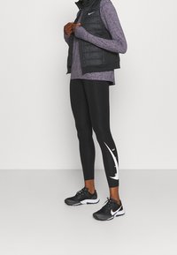 Černá prošívaná vesta přes fialové tričko s dlouhým rukávem, kombinovaná s černými legínami s bílým motivem a černými sportovními botami.