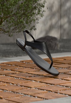 Μαύρο σανδάλι με λουράκι τύπου σλιπ και διάτρητο επάνω μέρος, παρουσιάζεται να αιωρείται πάνω από ξύλινη επιφάνεια με υφή από βότσαλα σε υπαίθριο χώρο.