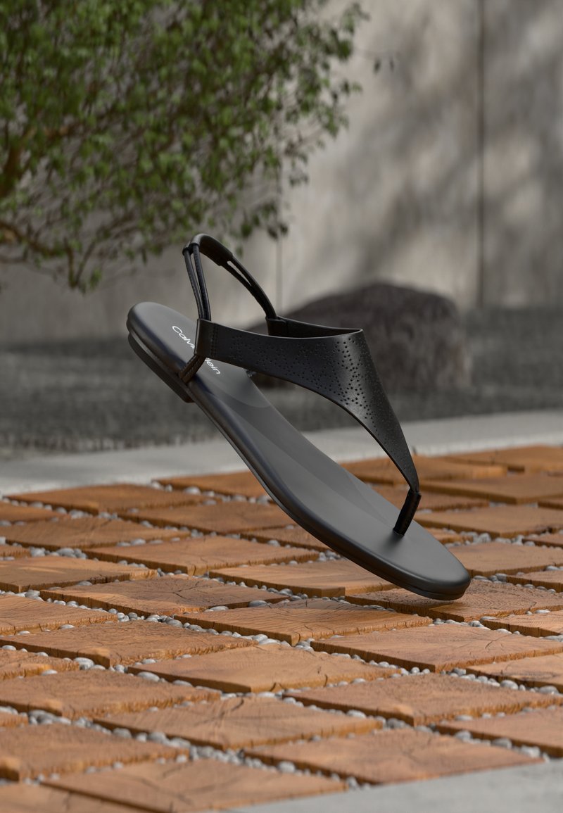Μαύρο σανδάλι με λουράκι τύπου σλιπ και διάτρητο επάνω μέρος, παρουσιάζεται να αιωρείται πάνω από ξύλινη επιφάνεια με υφή από βότσαλα σε υπαίθριο χώρο.