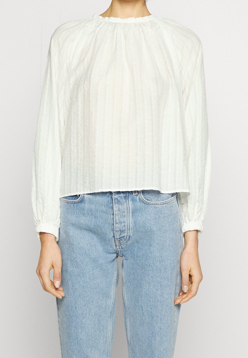 Personne portant un chemisier blanc texturé à manches longues avec des poignets élastiques, associé à un jean taille haute bleu clair, montré du cou aux hanches.