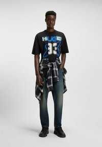 T-shirt grafica nera con scritte blu e bianche, abbinata a jeans azzurri chiari e a una camicia a quadri annodata in vita. Scarpe nere.