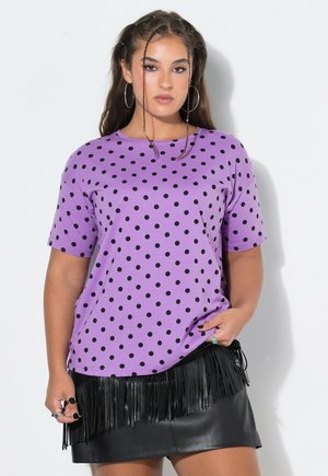 Donna con trecce lunghe che indossa una camicia viola a pois e una gonna di pelle nera con frange, in piedi contro un muro bianco.