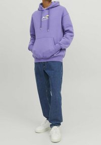 Felpa viola con tasca a marsupio, cordini e logo di montagna; abbinata a jeans blu e sneakers bianche. Texture morbida.