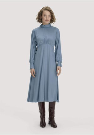 Vrouw met krullend haar, gekleed in een lichtblauwe jurk met lange mouwen en een hoge hals, en donkerbruine kniehoge laarzen, staand op een effen achtergrond.