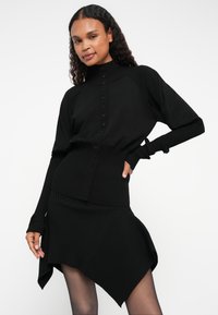 Suéter malha canelada preto e saia assimétrica com gola alta e fecho de botões. Apresenta mangas longas com buracos para os polegares.