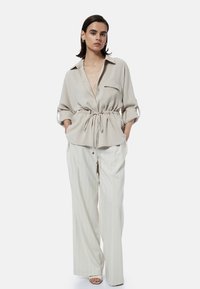Beige knoopshirt met een trekkoord in de taille, gecombineerd met witte broek met streepjes en minimalistische schoenen.