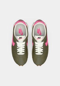 Olīvu zaļi sporta apavi ar rozā akcentiem, baltām auklām, strukturētu virsu un gumijas zoli. Nike logo redzams uz mēles un papēža.